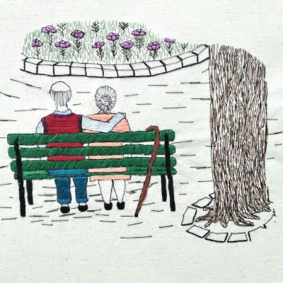 Bordado de casal idoso sentado em banco verde junto a árvore e flores roxas