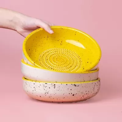 Três pratos fundos cerâmicos, amarelo e bege com manchas, empilhados sobre fundo rosa, com mão segurando o prato amarelo.