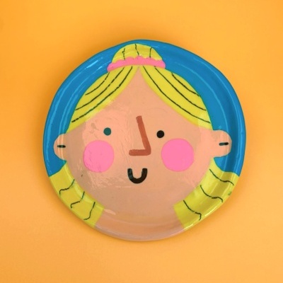 Prato decorativo redondo com desenho de rosto de criança em fundo azul e amarelo sobre superfície laranja