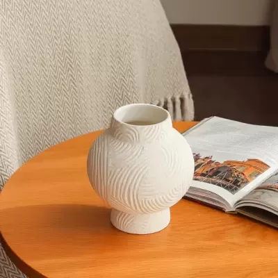 Vaso branco decorativo com relevo sobre mesa de madeira com revista aberta ao lado