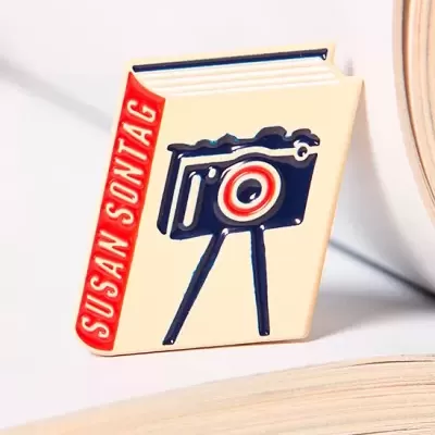 Broche esmalte livro com texto SUSAN SONTAG e desenho de máquina fotográfica