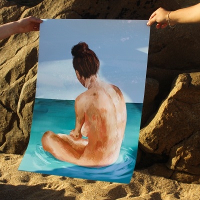 Poster com pintura de mulher nua sentada na água, segurado por duas mãos na praia