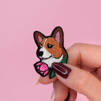 Broche bordado de cabeça de cão colorido segurado por mão com unhas vermelhas em fundo rosa