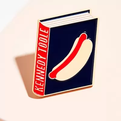 Pin metálico em forma de livro com texto 'KENNEDY TOOLE' e imagem de salsicha