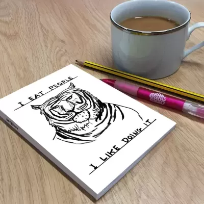 Caderno com ilustração de tigre e texto humorístico, caneta cor-de-rosa, lápis e chávena de café sobre mesa de madeira