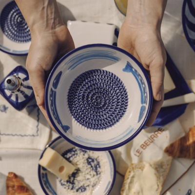 Tigela cerâmica branca e azul segurada por mãos sobre mesa com queijo, pão e louça azul