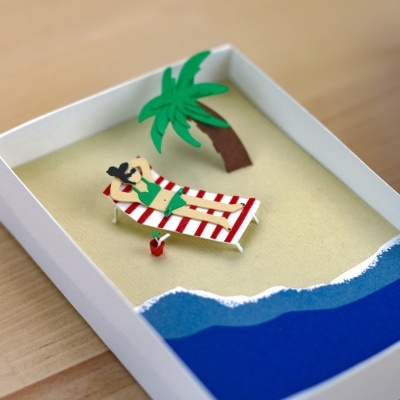 Miniatura de praia com pessoa em espreguiçadeira e palmeira numa caixa branca.