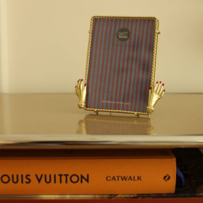 Suporte dourado em forma de mãos a segurar quadro riscado com logo, sobre mesa brilhante, com livros LOUIS VUITTON CATWALK e TOMMY NELSON em baixo.