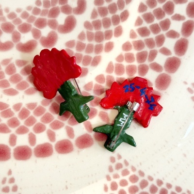 Duas broches em forma de flor vermelha com caules verdes e texto azul e branco, sobre fundo branco com padrão vermelho.