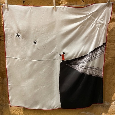 Lenço branco com desenho de pessoa e pássaros, pendurado em parede de pedra