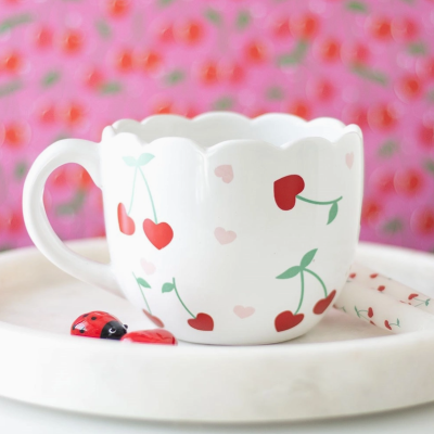 Caneca branca decorada com cerejas vermelhas e corações pequenos, numa mesa branca com fundo rosa floral