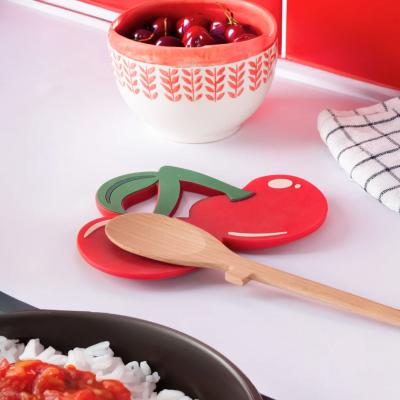 Base para colher em forma de cereja vermelha com folhas verdes em cozinha branca com tigela cerâmica e pano