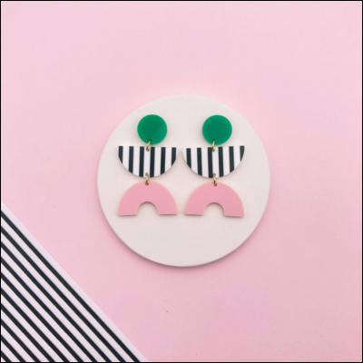 Brincos de resina coloridos em fundo rosa com círculo branco e papel listrado.