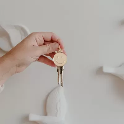 Chaveiro dourado com rosto sorridente segurado por mão contra fundo cinza.