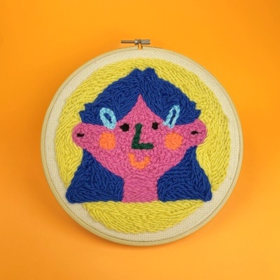 Bordado em bastidor com rosto estilizado colorido sobre fundo laranja