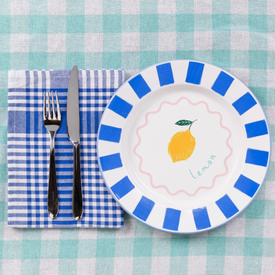 Prato decorado com faixas azuis e limão, talheres sobre guardanapo azul e branco em mesa xadrez verde claro