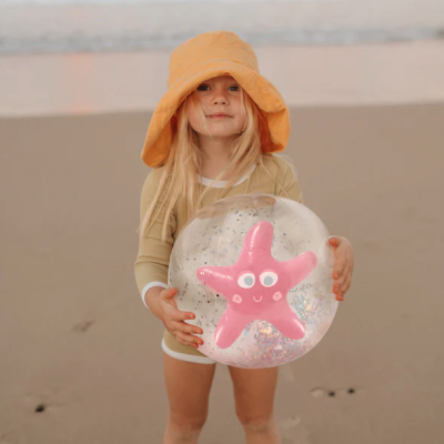 Criança com chapéu amarelo segurando bola transparente com estrela do mar rosa na praia