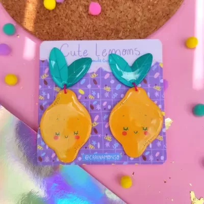 Brincos amarelos em forma de limão com folhas verdes sobre cartão roxo decorado, em fundo rosa com confetes.