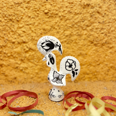 Estatueta de galo branca com desenhos florais a preto sobre superfície amarelo texturado com fitas coloridas