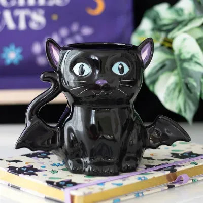 Caneca preta em forma de gato com asas de morcego sobre um caderno com desenhos de gatos.