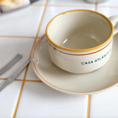 Xícara de cerâmica creme com pires e talheres sobre azulejos brancos