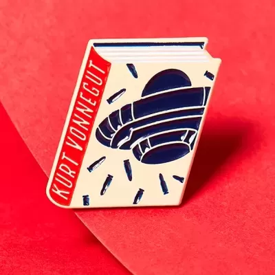 Pino esmaltado em forma de livro com título KURT VONNEGUT e ilustração azul, sobre fundo vermelho.