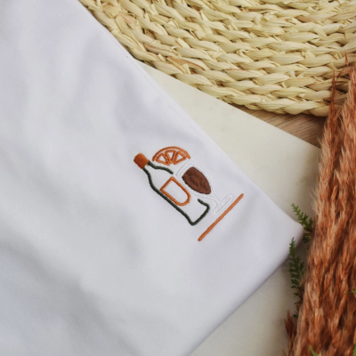 T-shirt branca com bordado de garrafa, fatia de laranja e copo, sobre fundo natural e plantas secas