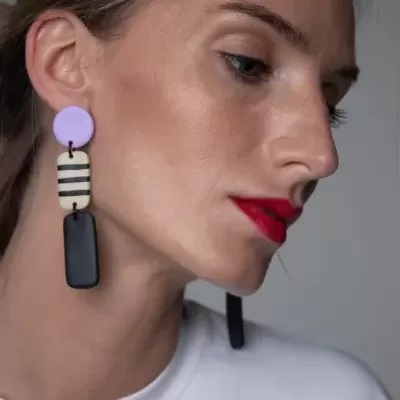 Brincos pendentes coloridos com formas geométricas usados por mulher com batom vermelho e camiseta branca.