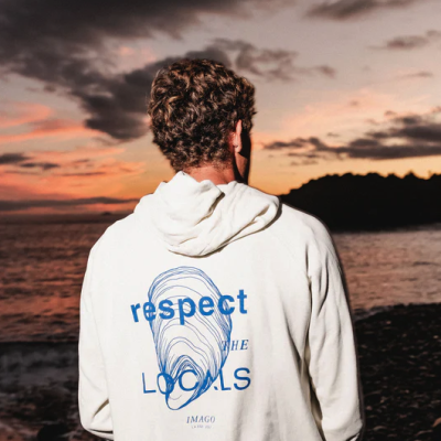 Pessoa de costas com hoodie branco e estampa azul de texto e desenho circular, praia ao pôr do sol ao fundo