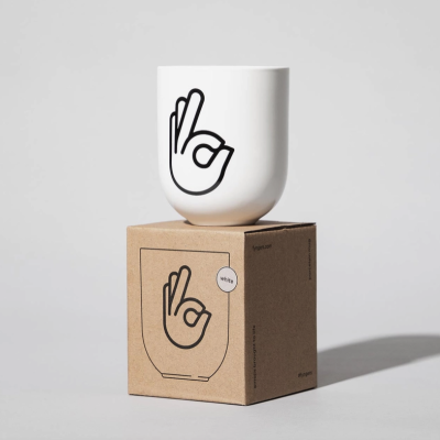 Caneca branca com símbolo mão OK em preto sobre caixa castanha.
