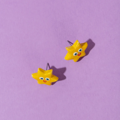 Brincos em forma de estrela amarelas com rosto desenhado em fundo roxo