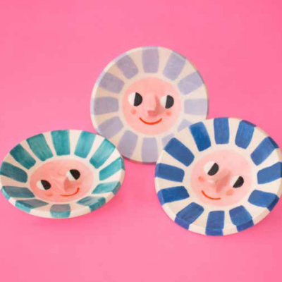 Três pratos decorativos redondos com faces sorridentes e riscas coloridas em fundo cor-de-rosa