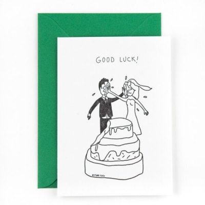 Cartão branco com ilustração a preto de noiva a beijar noivo sobre bolo, texto GOOD LUCK!, com envelope verde
