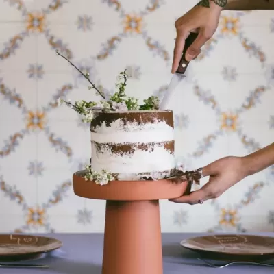 Bolo rústico com flores num suporte terracota sendo cortado por uma pessoa com tatuagem.