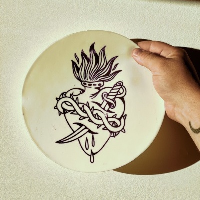 Prato redondo branco com desenho preto de coração flamejante, faca e vinha espinhosa, segurado por mão