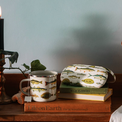 Caneca e estojo com estampa de peixes sobre livros numa mesa de madeira com vela acesa ao lado