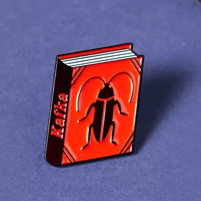 Pin metálico em forma de livro vermelho com dibujo de inseto e texto Kafka