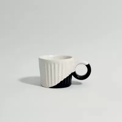 Xícara pequena de cerâmica branca e preta, com cabo circular preto e branco, sobre fundo cinza