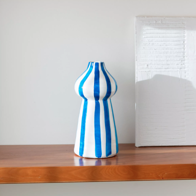 Vaso cerâmico azul e branco com riscas verticais numa prateleira de madeira frente a parede branca