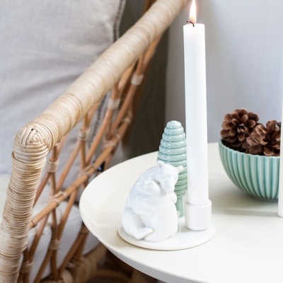 Mesa redonda branca com castiçal, figura de urso, objeto em forma de árvore, bowl com pinecones e cadeira de vime com almofada