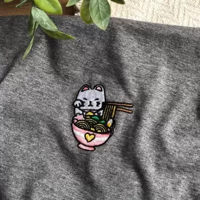 Bordado de gato cinzento numa tigela rosa com noodles num tecido cinzento
