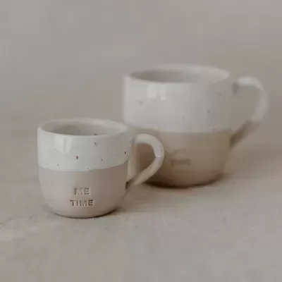Dois copos de cerâmica branco e bege com a inscrição ME TIME em fundo neutro