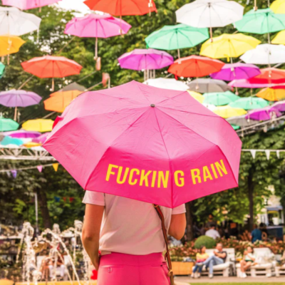 Guarda-chuva rosa com texto amarelo em ambiente exterior com muitos guarda-chuvas coloridos suspensos