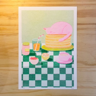 Ilustração de gato rosa em cima de panquecas numa mesa com toalha xadrez e utensílios de pequeno-almoço