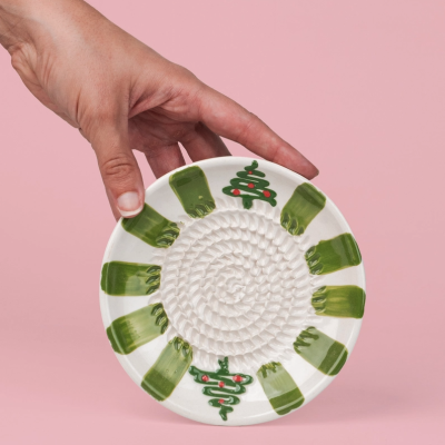 Prato de cerâmica branco decorado com formas verdes e árvores de Natal, contra fundo rosa.