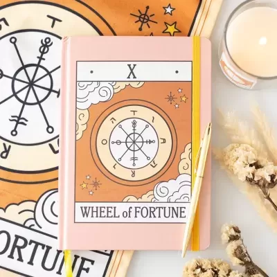 Caderno rosa com design tarot e caneta dourada, acompanhado de vela e flores secas