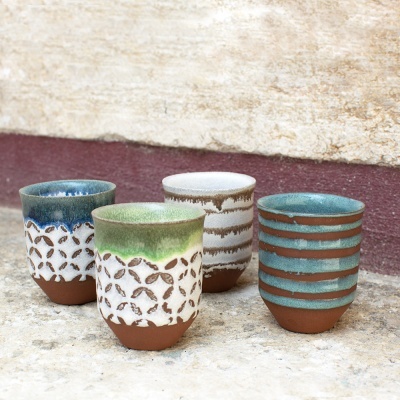 Quatro copos de cerâmica esmaltados com padrões geométricos, dispostos sobre pedra.