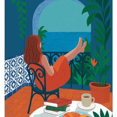 Mulher sentada numa varanda com comida, bebidas, livros e vista para o mar.