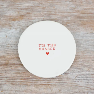 Placa decorativa branca com texto vermelho 'TIS THE SEASON' e coração vermelho sobre madeira clara