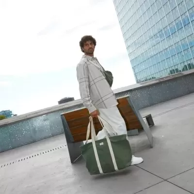 Homem segurando mala de lona verde ao lado de banco em área urbana